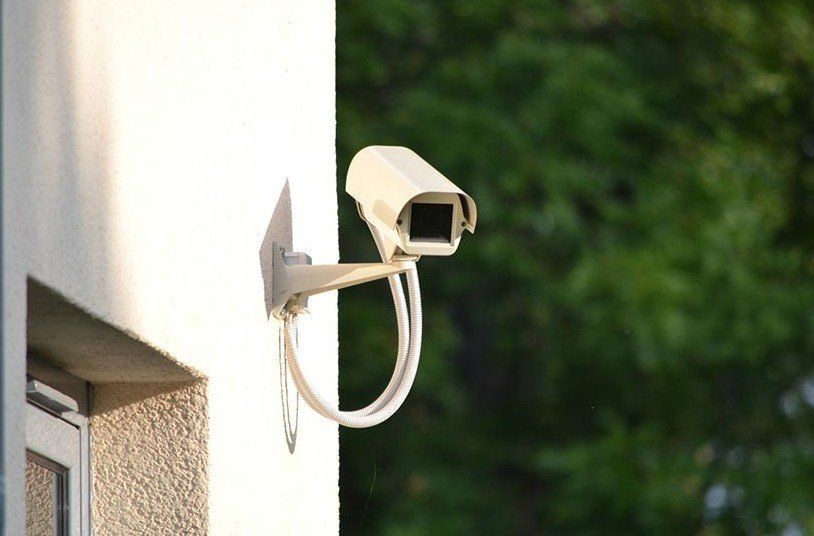 Security camera mounted on a white building exterior with a blurred green tree backdrop.