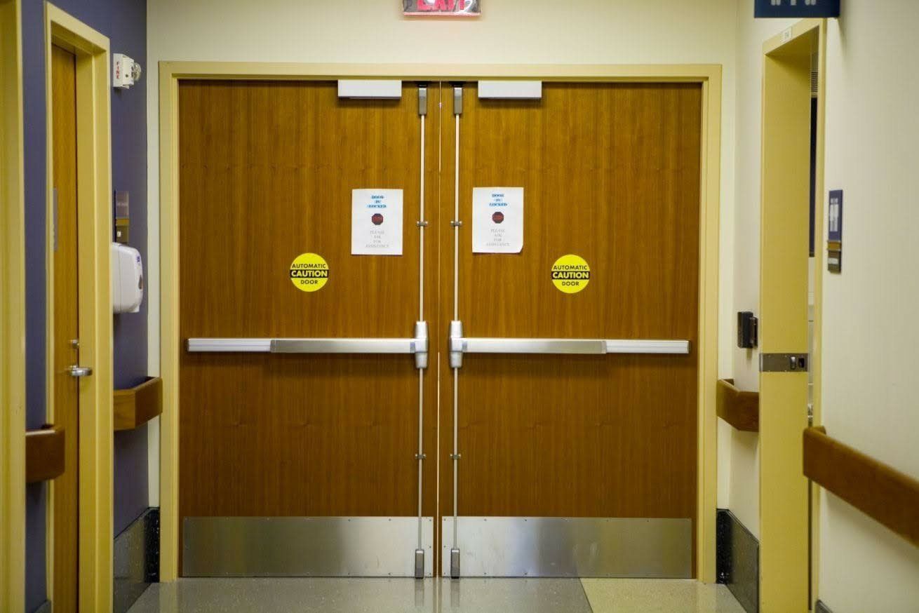 Double wooden doors with silver bars, automatic door stickers, and exit sign in a hallway.