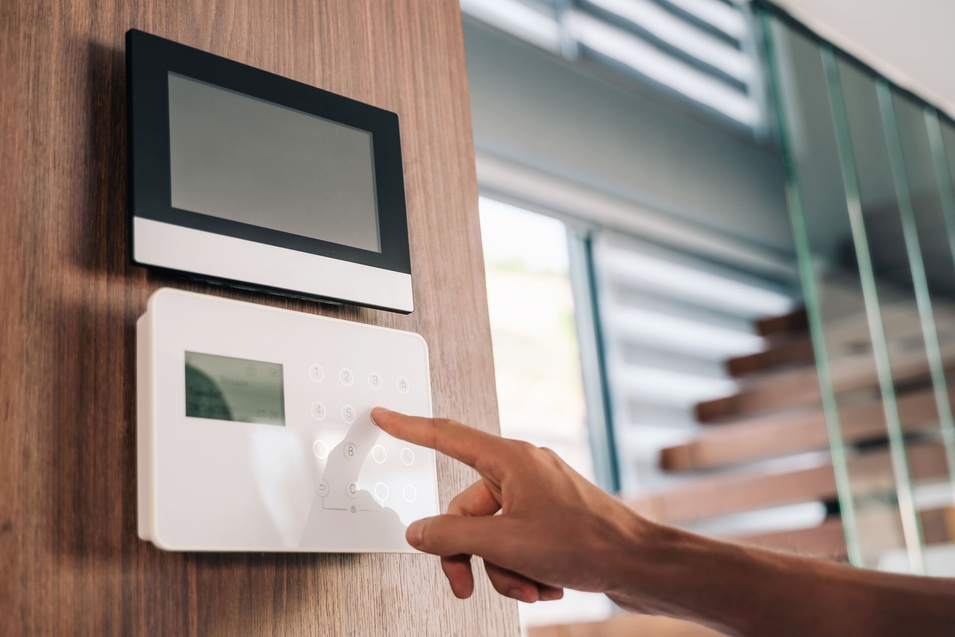 Hand pressing a button on a white security system keypad mounted on a wooden wall, with a black screen above.