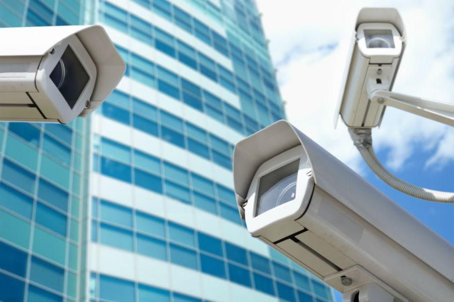 Three white security cameras point towards a blue-glass building. The cameras are set against a partly cloudy sky.