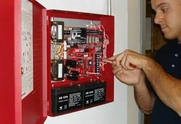 Man working on a red fire alarm control panel mounted on a wall, using a screwdriver. The panel's interior contains wiring, circuit boards, and batteries.