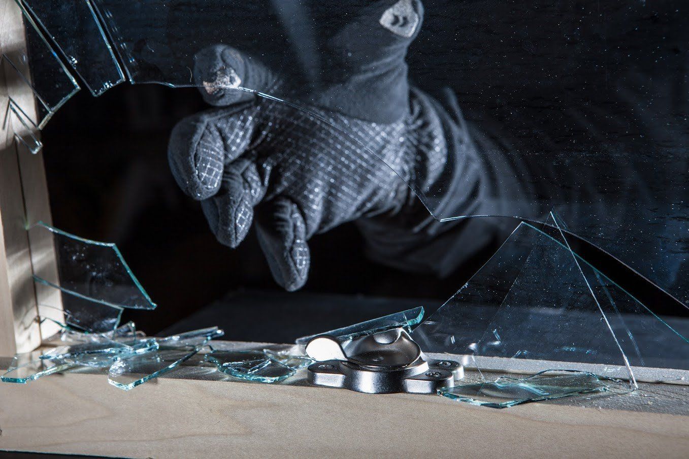 Gloved hand reaching through a shattered window, likely committing a burglary. Dark setting, broken glass.