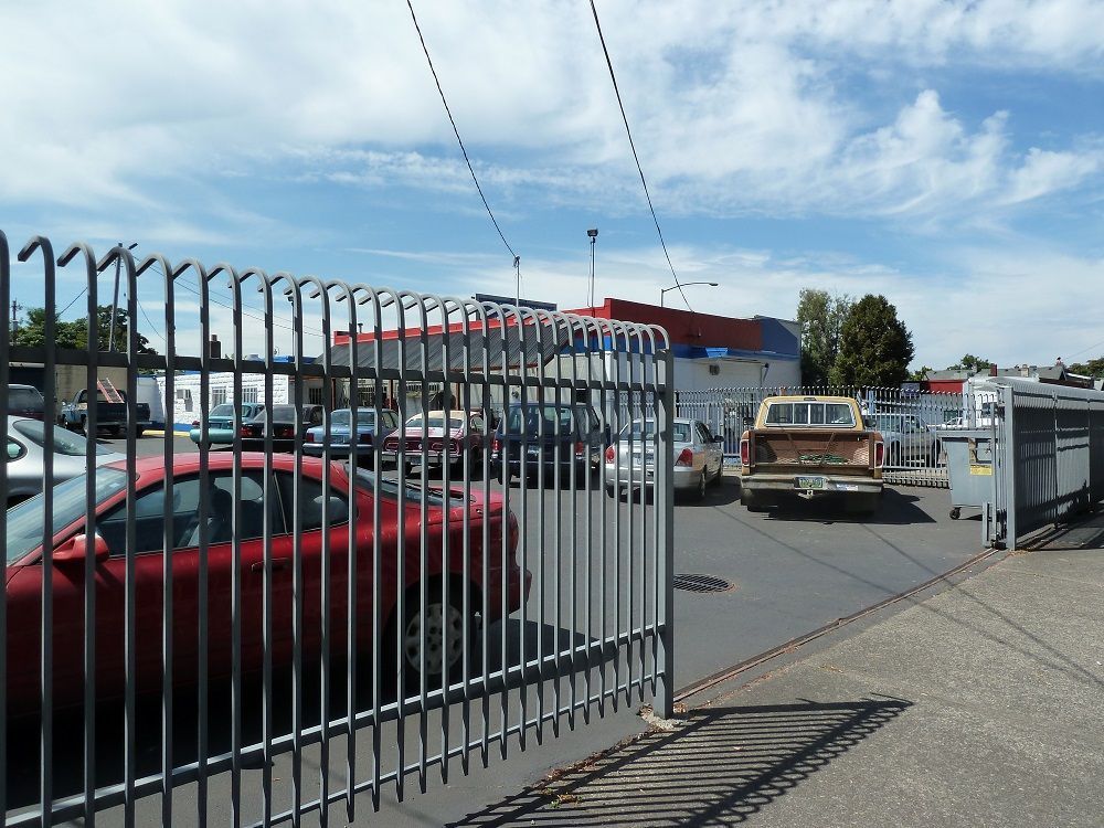 A red car and a brown pickup truck are parked behind a metal fence on a sunny day. Buildings and other cars are visible in the background.