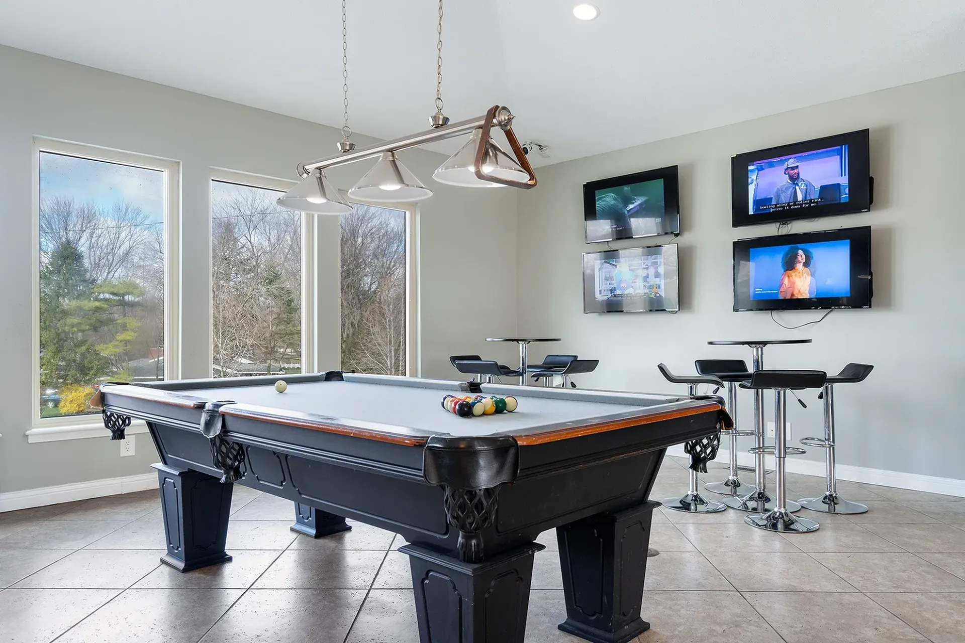 Pool table in a game room with bar tables, multiple TVs, and a view of trees through large windows.