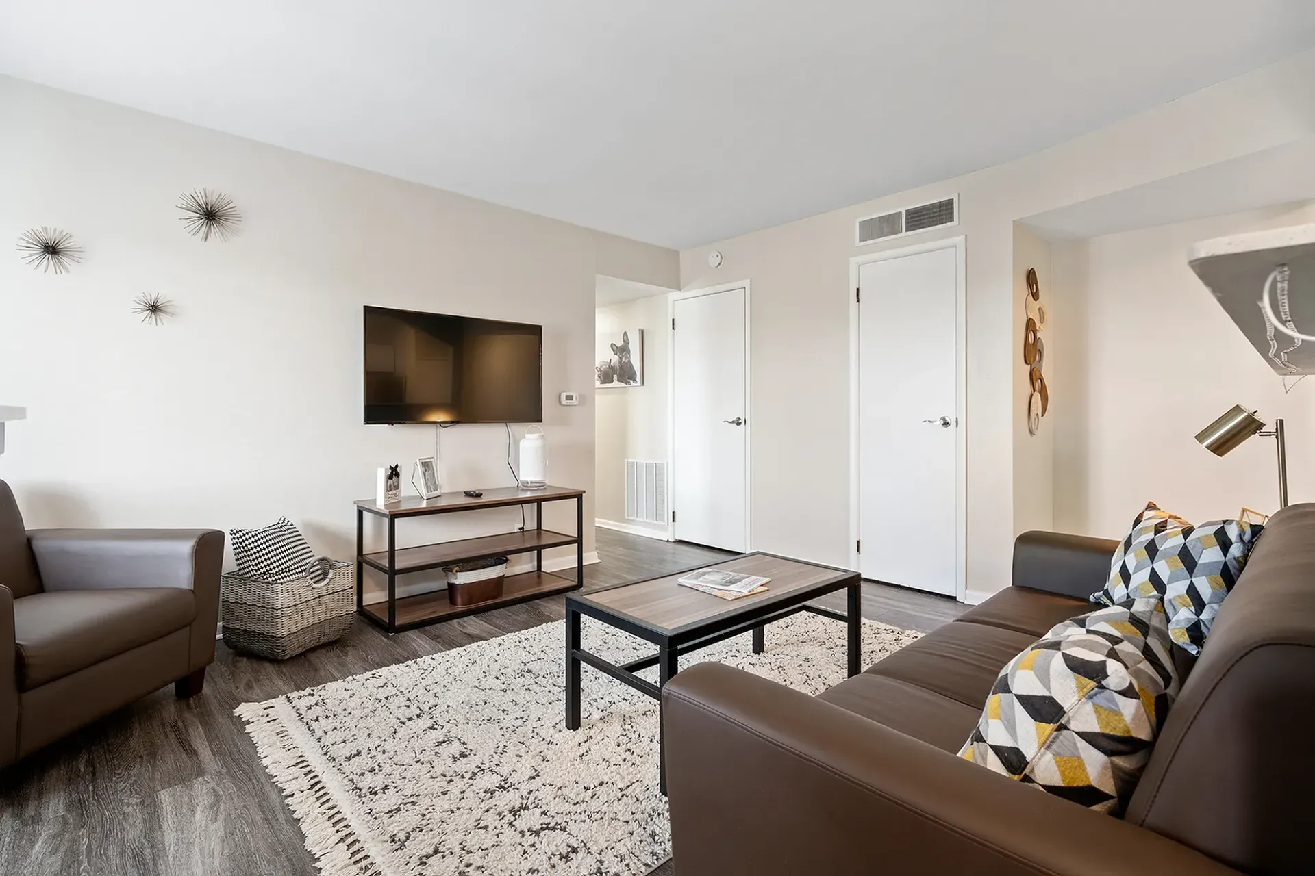 Living room with brown sofa, gray armchair, TV, and rug. Light walls and wood flooring.