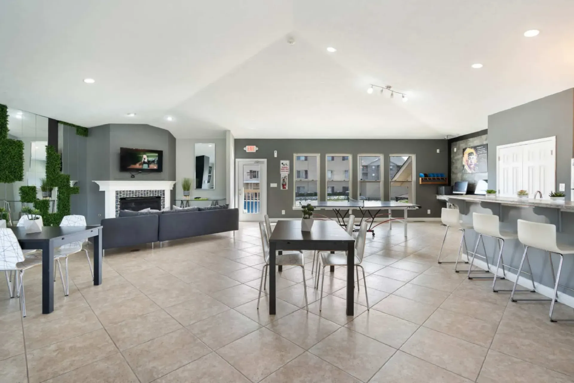Open-concept community lounge with tables, bar, seating, and fireplace against gray walls and tile flooring.