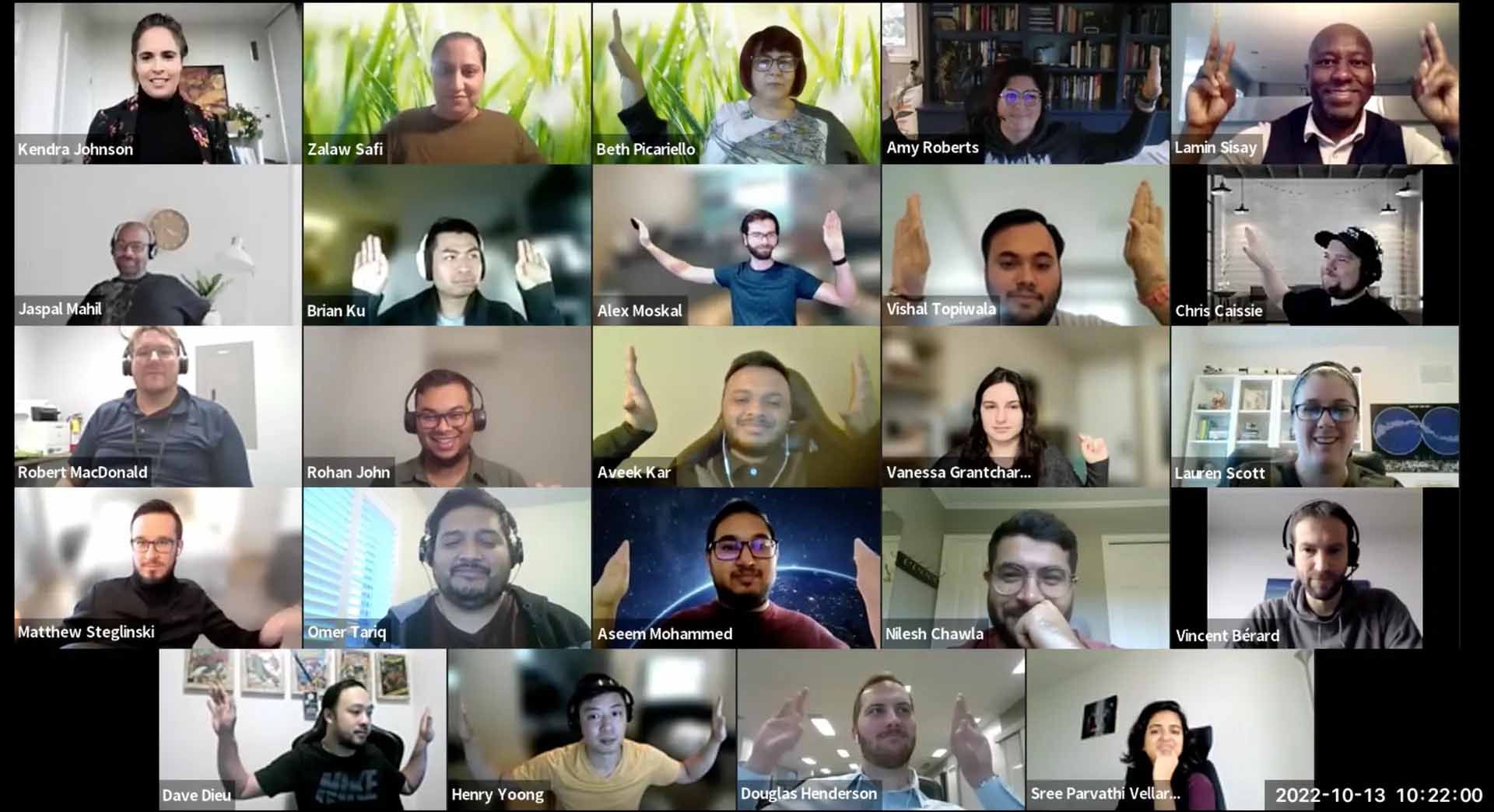 Group of people in a video call raising hands, many smiling, against various backgrounds, indoors.