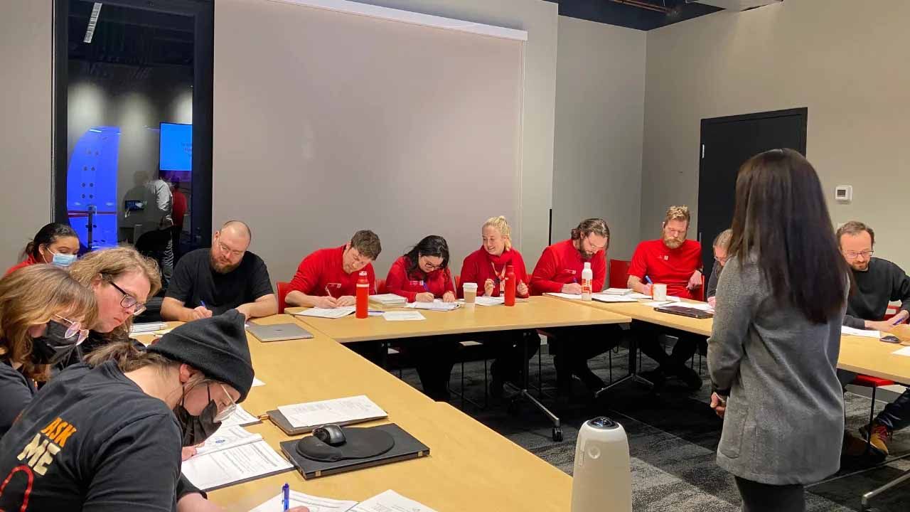 People seated around a U-shaped table writing. One person faces them, speaking. Red shirts, papers, a room setting.