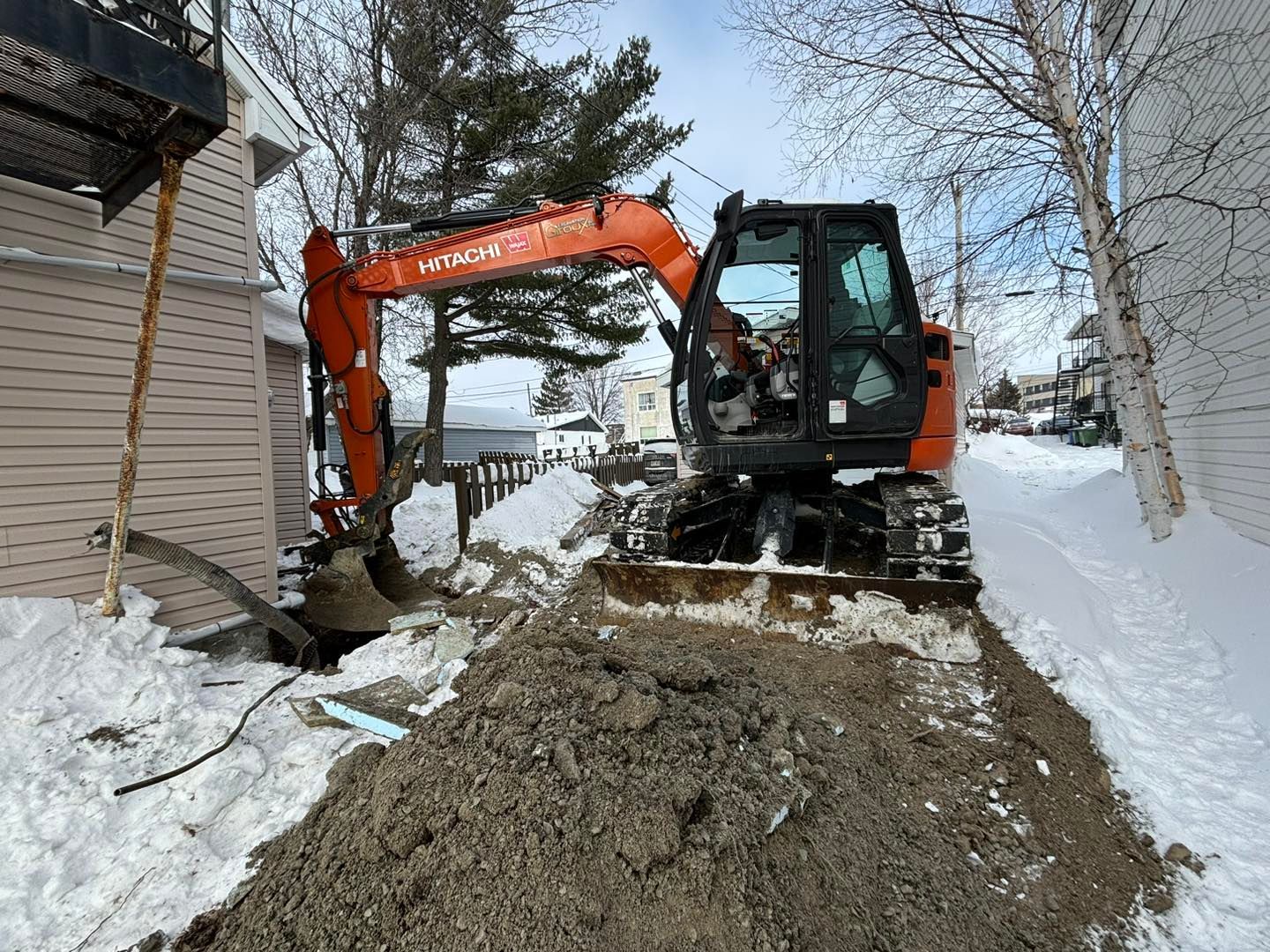 Une excavatrice creuse un trou dans la neige devant une maison.