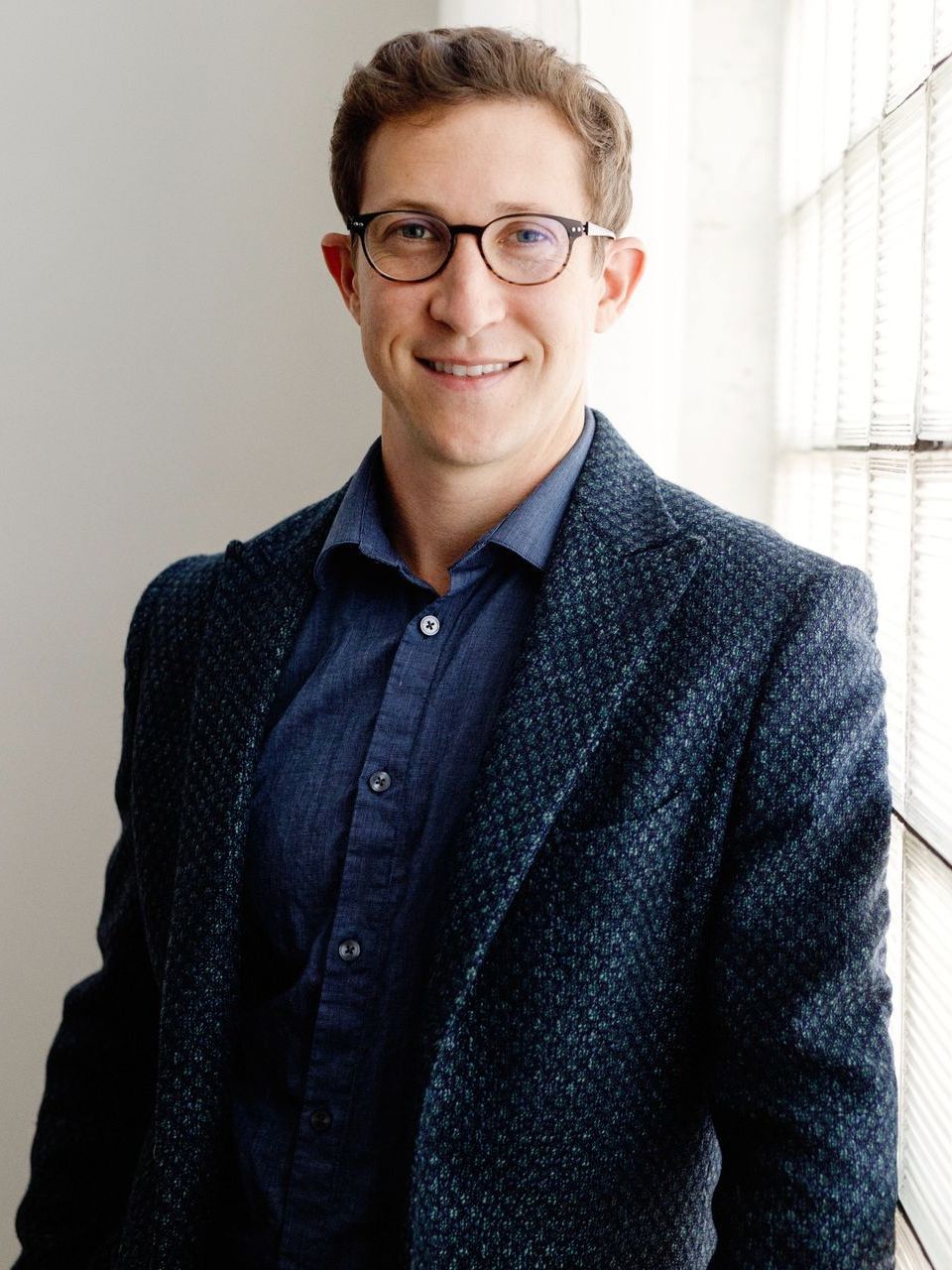 Man in glasses, smiling, wearing a blue shirt and dark textured jacket, standing near a window.