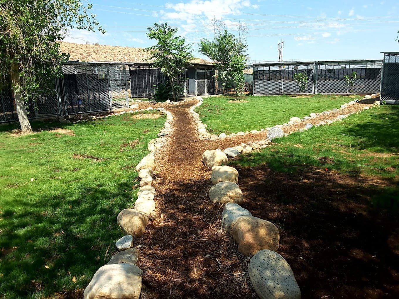 Stone-lined dirt path leading to a farmhouse courtyard with green grass and fenced buildings under a blue sky