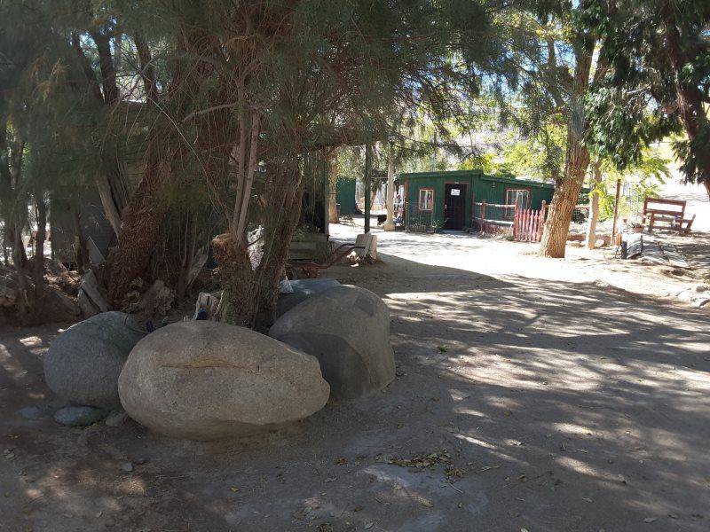 Shady dirt path with large rocks and trees leading to a small green building