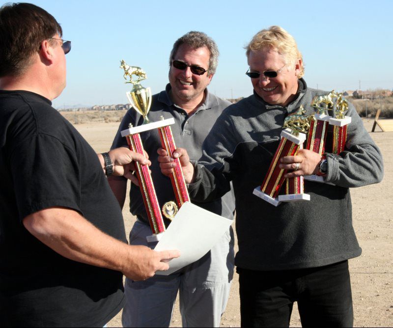 Three men outdoors, two holding trophies and smiling while a man hands them papers