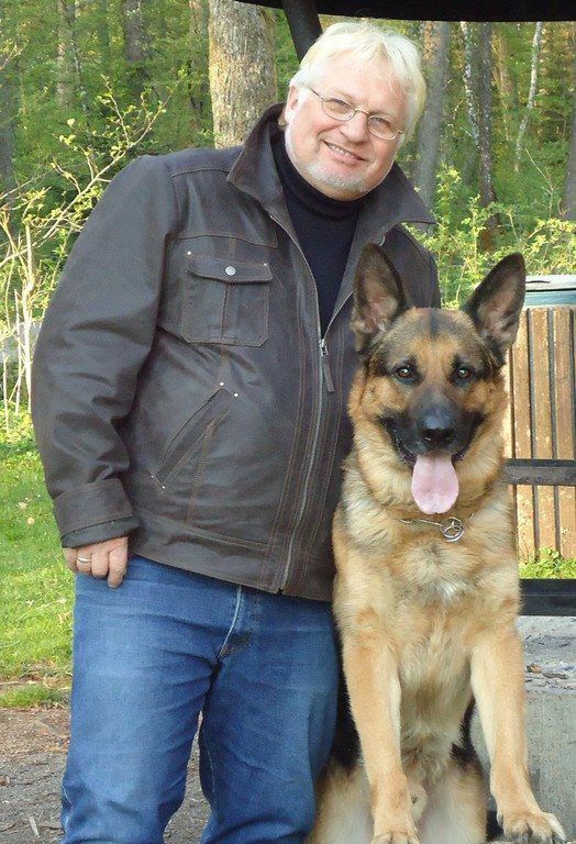 Man standing beside a German Shepherd outdoors, both facing the camera, with trees and a railing behind them.
