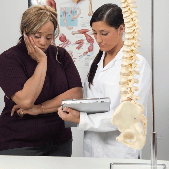 Woman looking concerned as chiropractor points to spinal model, holding clipboard