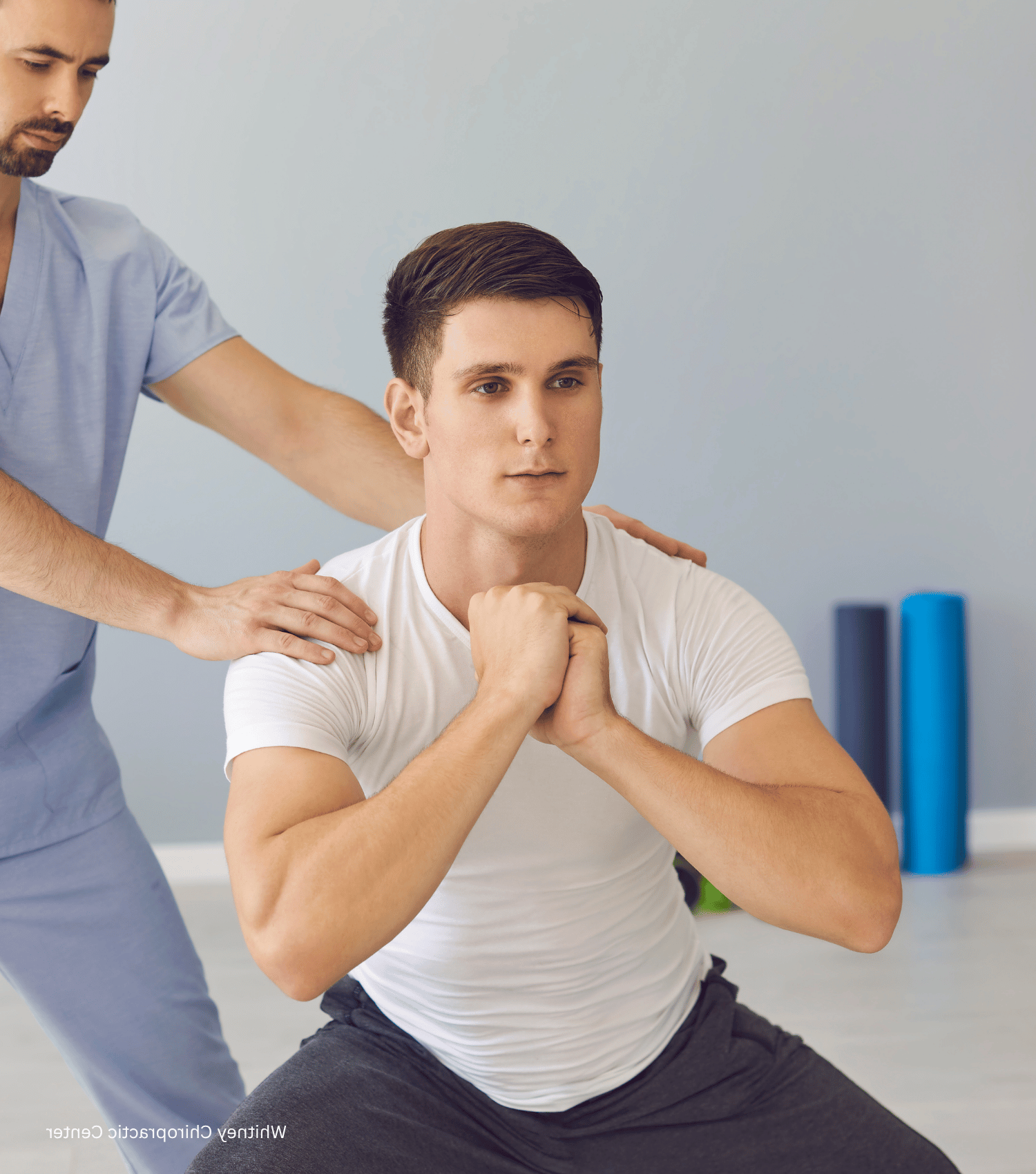 A man squats with guidance from a medical professional. They are in a clinic; blue and gray colors.