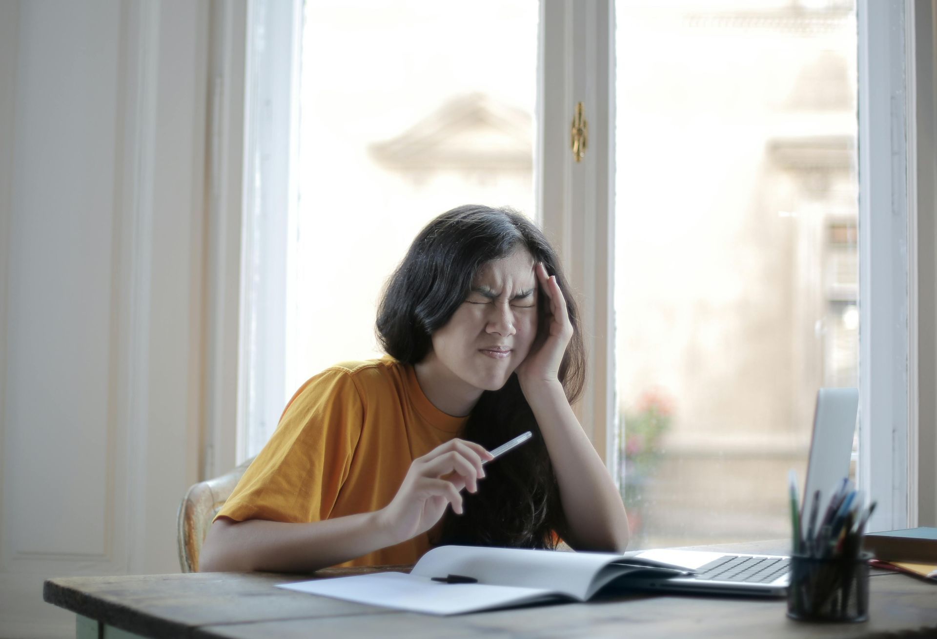A young woman with a headache, sitting at a desk with a laptop, notepad, and pen near a window.