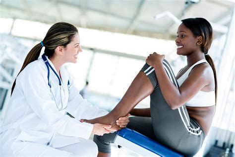 Doctor examining athlete's ankle; indoors, both smiling, one in sportswear, the other in a lab coat.