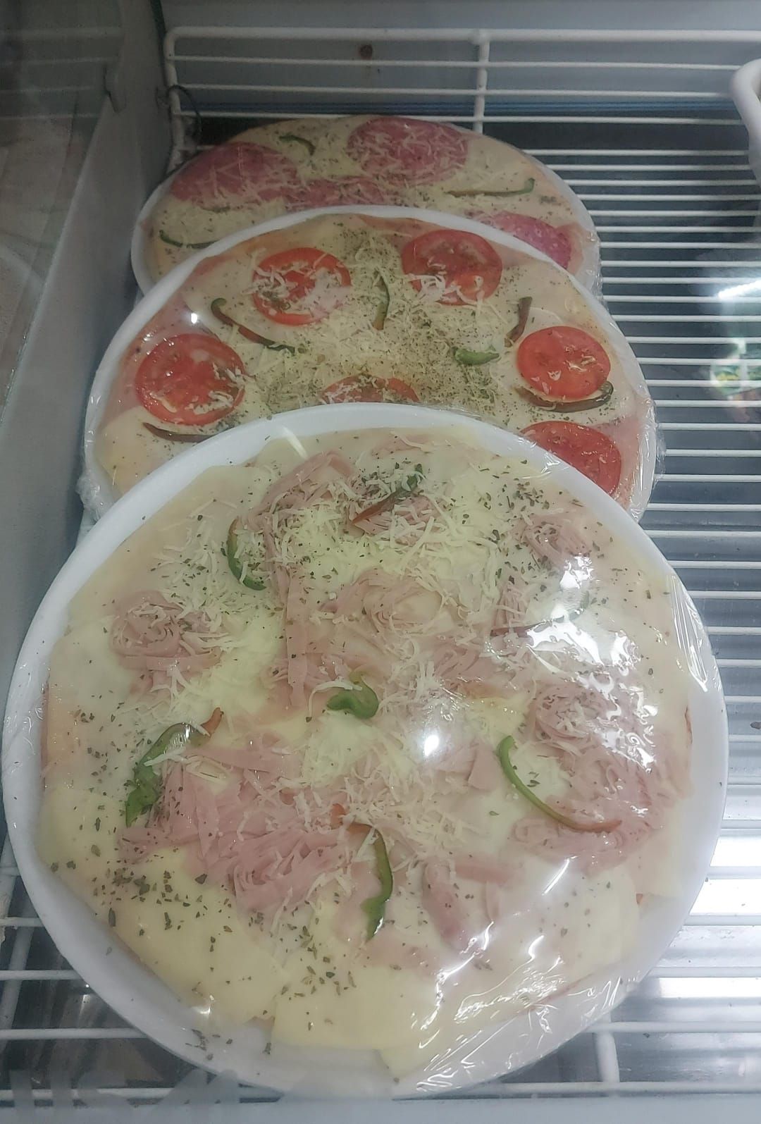 Tres pizzas envueltas con distintos ingredientes, expuestas en un estante dentro de una vitrina refrigerada.