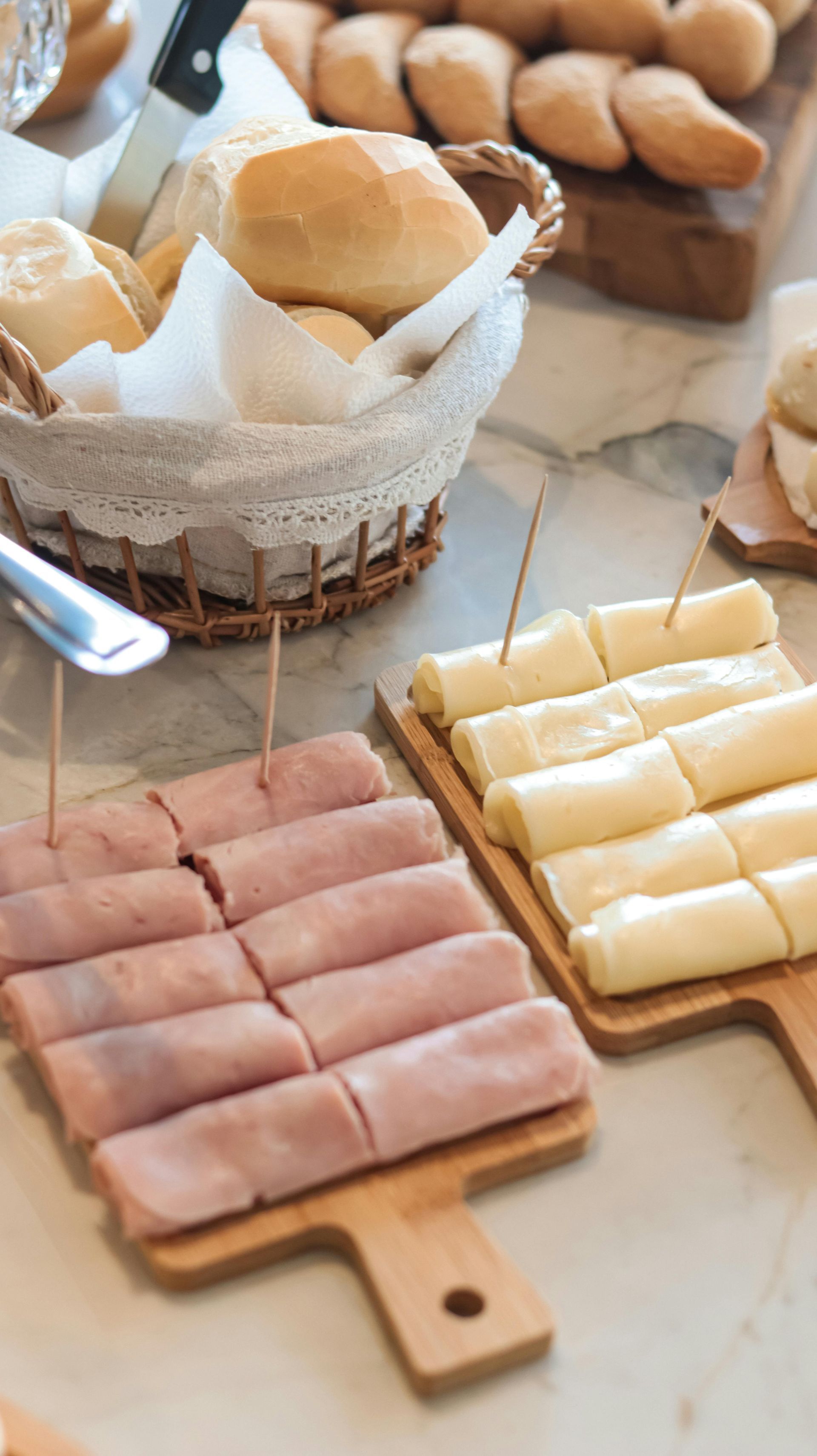 Pan y rollitos de jamón y queso en lonchas sobre tablas de madera, una cesta de panecillos al fondo.