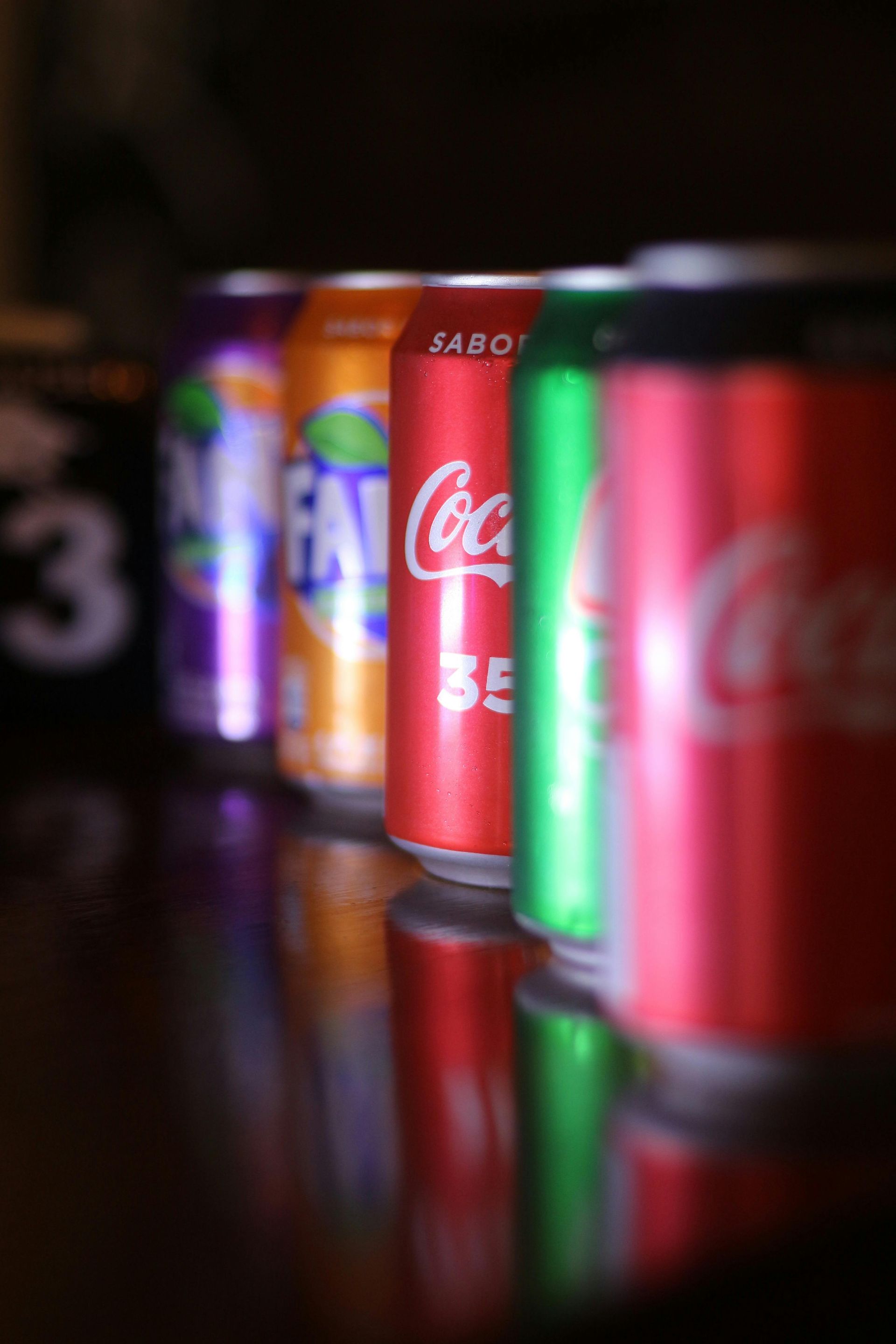 Cinco latas de refresco de colores en fila, incluida una Fanta morada y naranja, y una Coca-Cola roja, verde y roja.