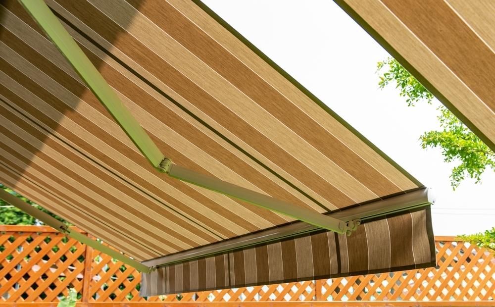 A Brown and Green Striped Awning is Hanging Over a Wooden Fence — Lismore Curtains & Blinds In Ballina, NSW