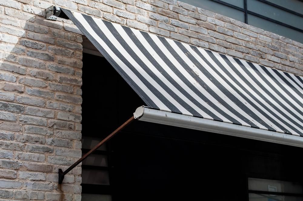A Black and White Striped Awning is Hanging From the Side of a Brick Building — Lismore Curtains & Blinds In Byron Bay, NSW
