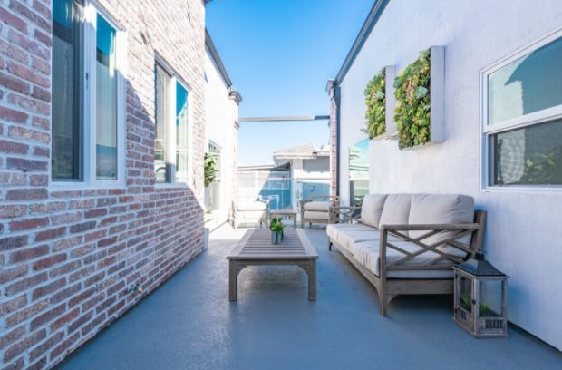 Bright rooftop patio with white and brick walls, gray sofa, coffee table, and potted greenery under blue sky