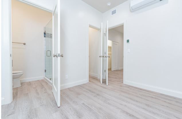 Bright empty bedroom with white walls, light wood floor, mirrored closet, and open doors to a bathroom and hall