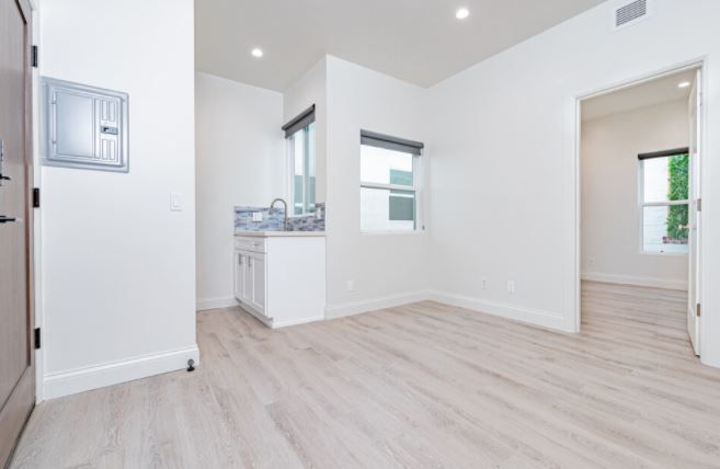 Bright empty apartment room with light wood floors, white walls, and a small kitchen opening.