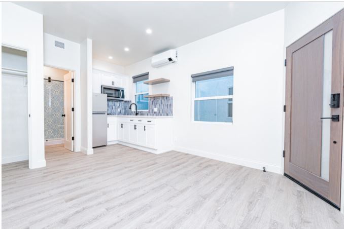 Bright empty modern room with white walls, light wood floors, kitchenette, windows, and a wooden door