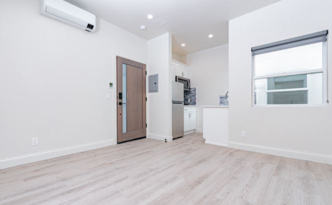 Empty modern studio apartment with white walls, light wood floors, air conditioner, and small kitchen area