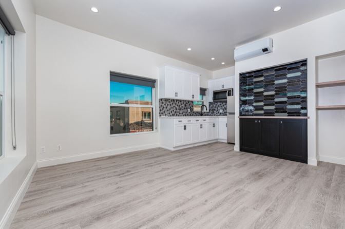 Bright modern empty room with light wood flooring, white walls, kitchenette, and large window.