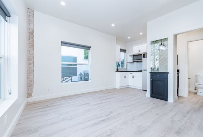 Bright, empty modern living room with light wood floors, white walls, and a small kitchen area.