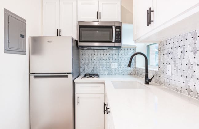 Bright white modern kitchen with stainless steel fridge, microwave, and black faucet over white counters.