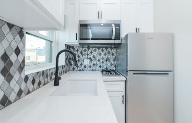 Modern white kitchen with gray tile backsplash, stainless fridge, black faucet, and white countertops