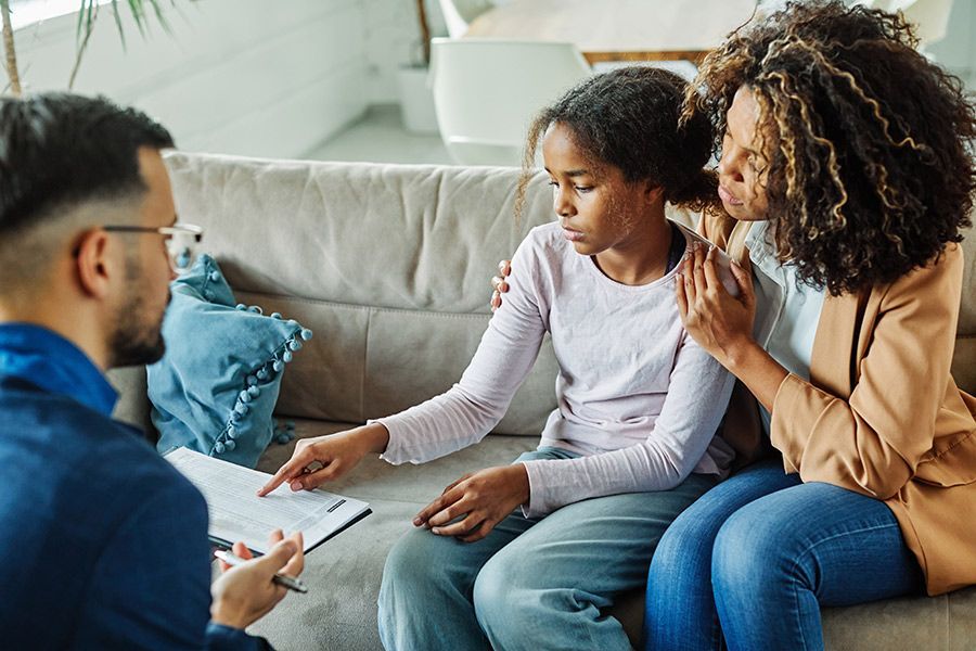 A Therapist Points to Paperwork While a Mother Comforts Her Daughter on a Sofa — Thrive Therapy Solutions in Mossman, QLD