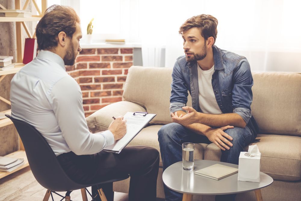 A Man is Sitting on a Couch Talking to a Psychologist — Thrive Therapy Solutions in Cairns City, QLD