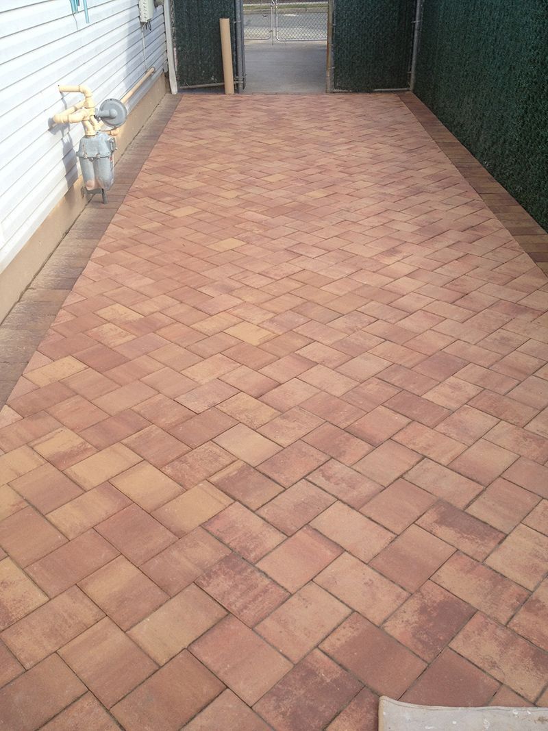 Brick driveway with herringbone pattern, leading to a gate, bordered by a green hedge and a white house.