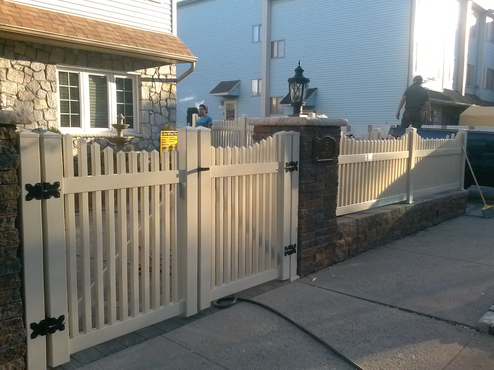 Beige picket fence with gate, bordering a brick wall and sidewalk. House visible in the background.