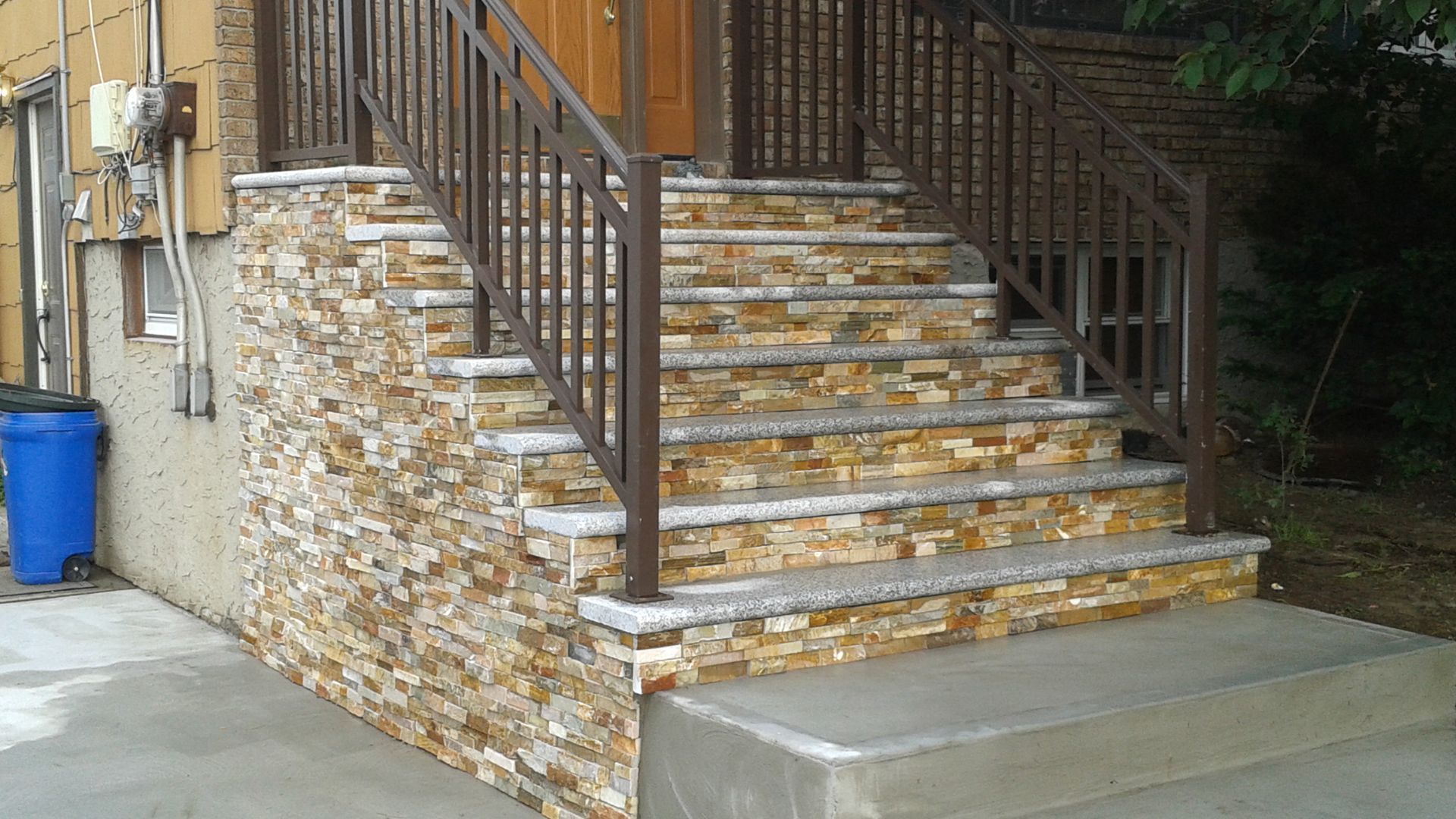 Stone-faced outdoor stairs with a brown metal railing leading to a door; a blue trash bin is visible.