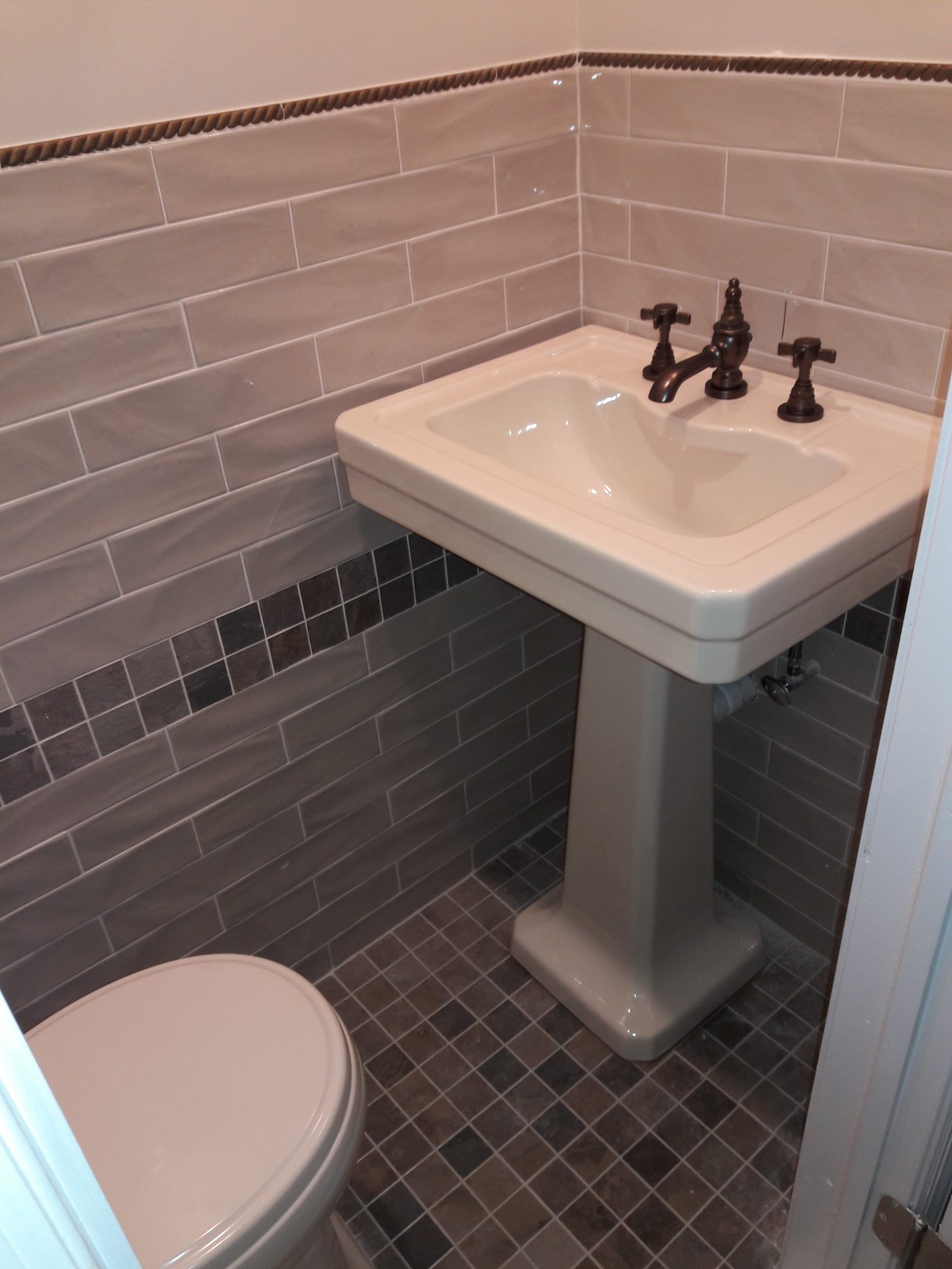 Bathroom with gray tiled walls and floor, pedestal sink, toilet, and dark faucet.