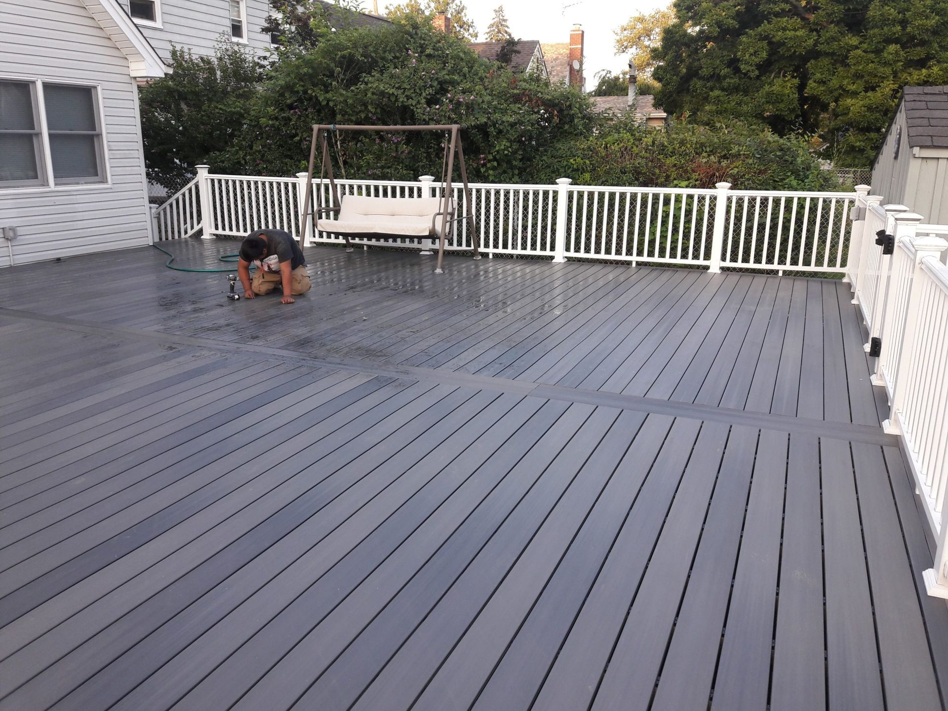 Person cleaning a gray deck with a hose; white railing surrounds the deck; trees in the background.