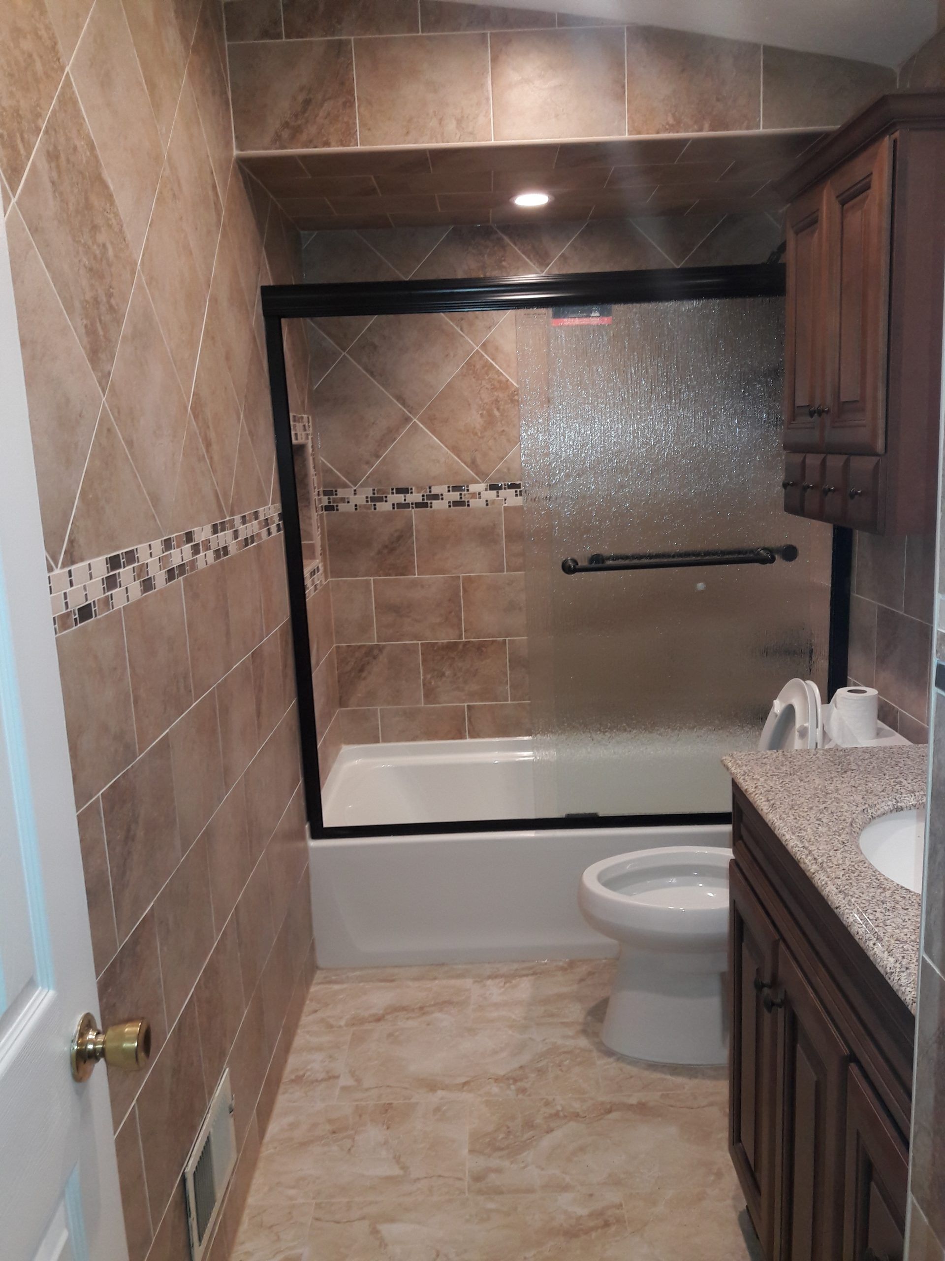 Bathroom with brown tiled walls, shower with glass door, white tub, toilet, and vanity.