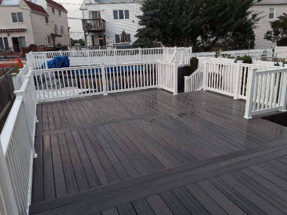 Grey deck with white railing surrounding a pool.  Buildings in the background.