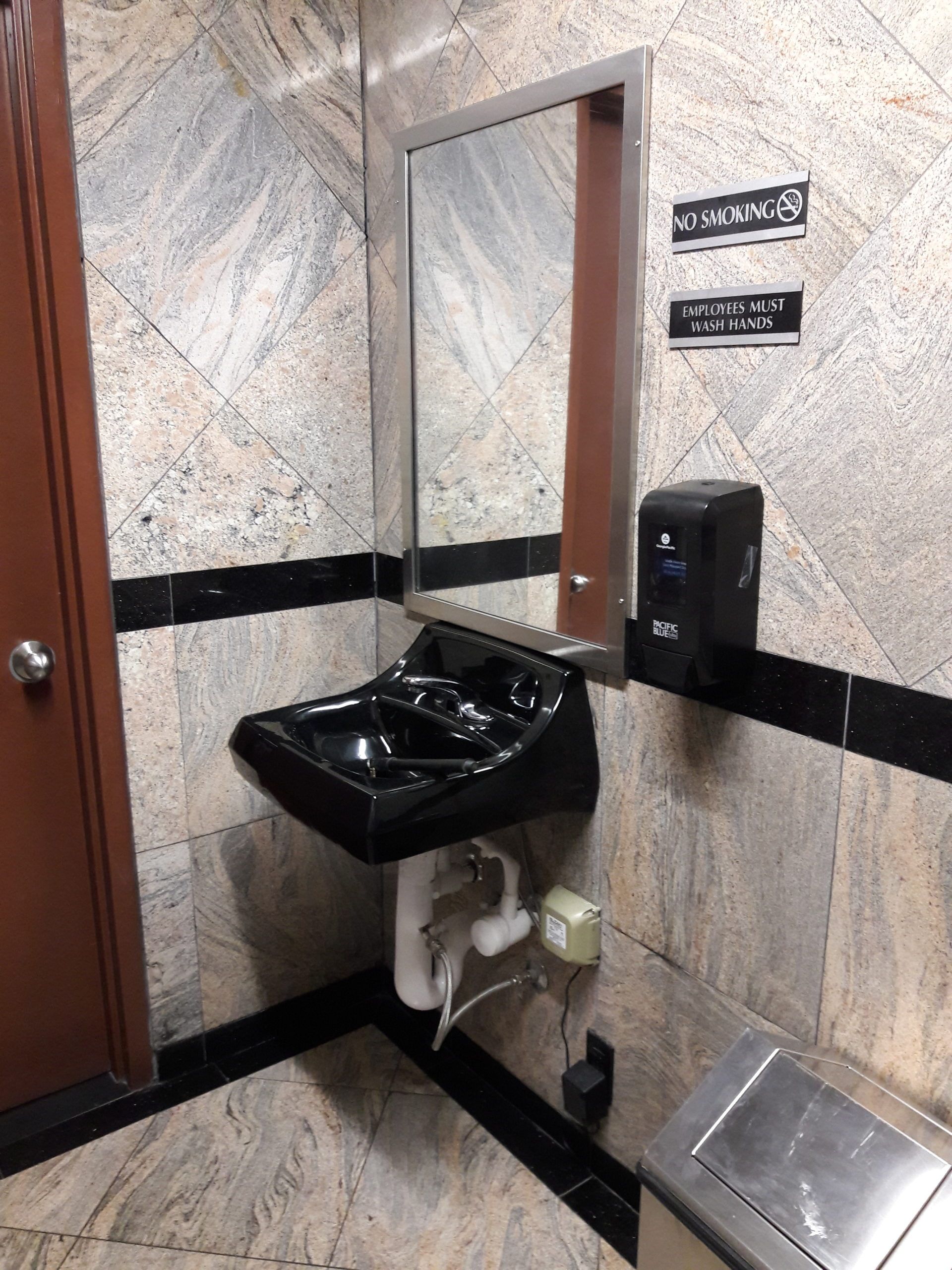 A black sink with a mirror and soap dispenser in a restroom with marble walls and a trash can.