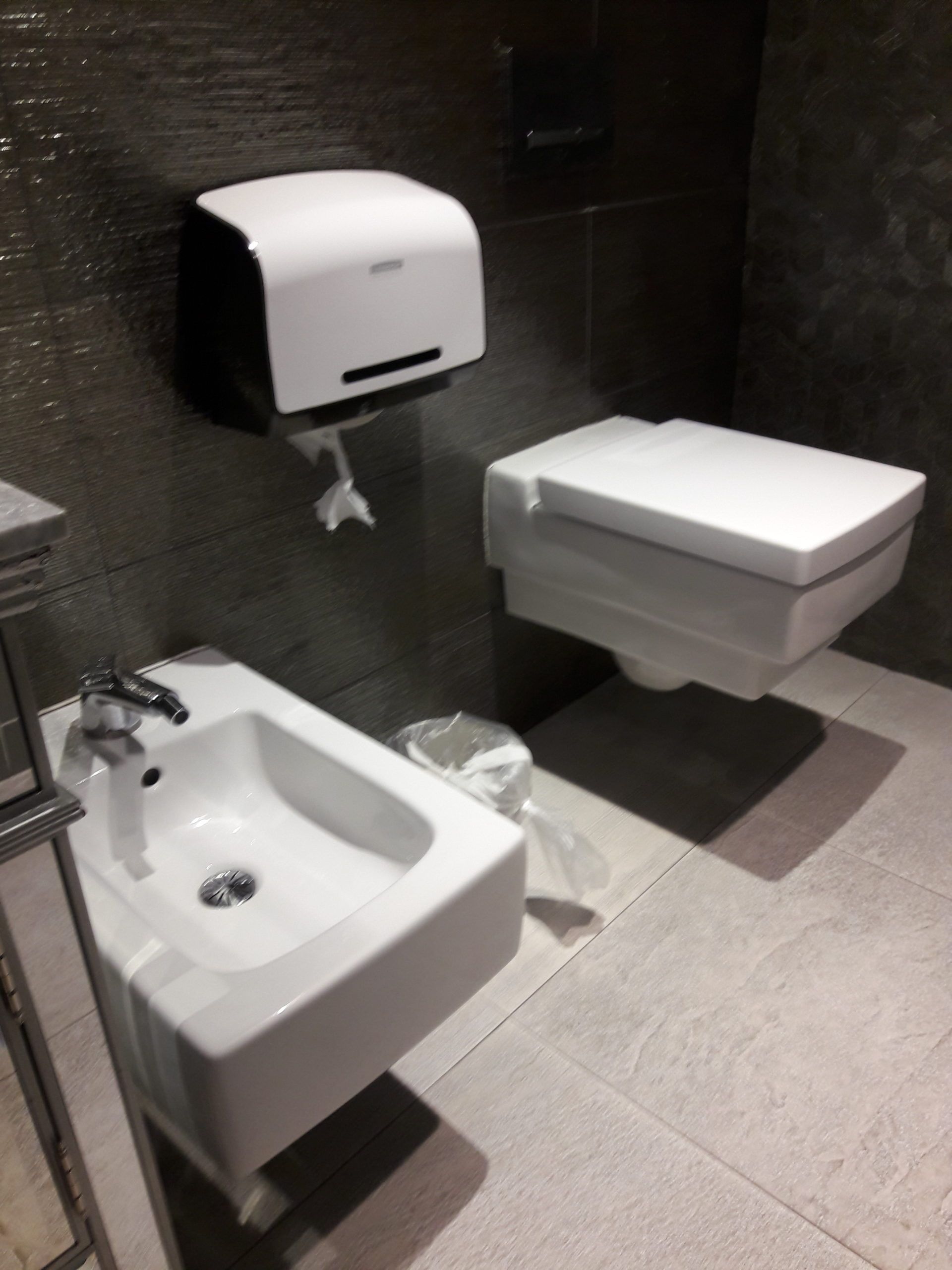 A modern bathroom with a bidet, wall-mounted toilet, and paper towel dispenser on a gray tiled wall.
