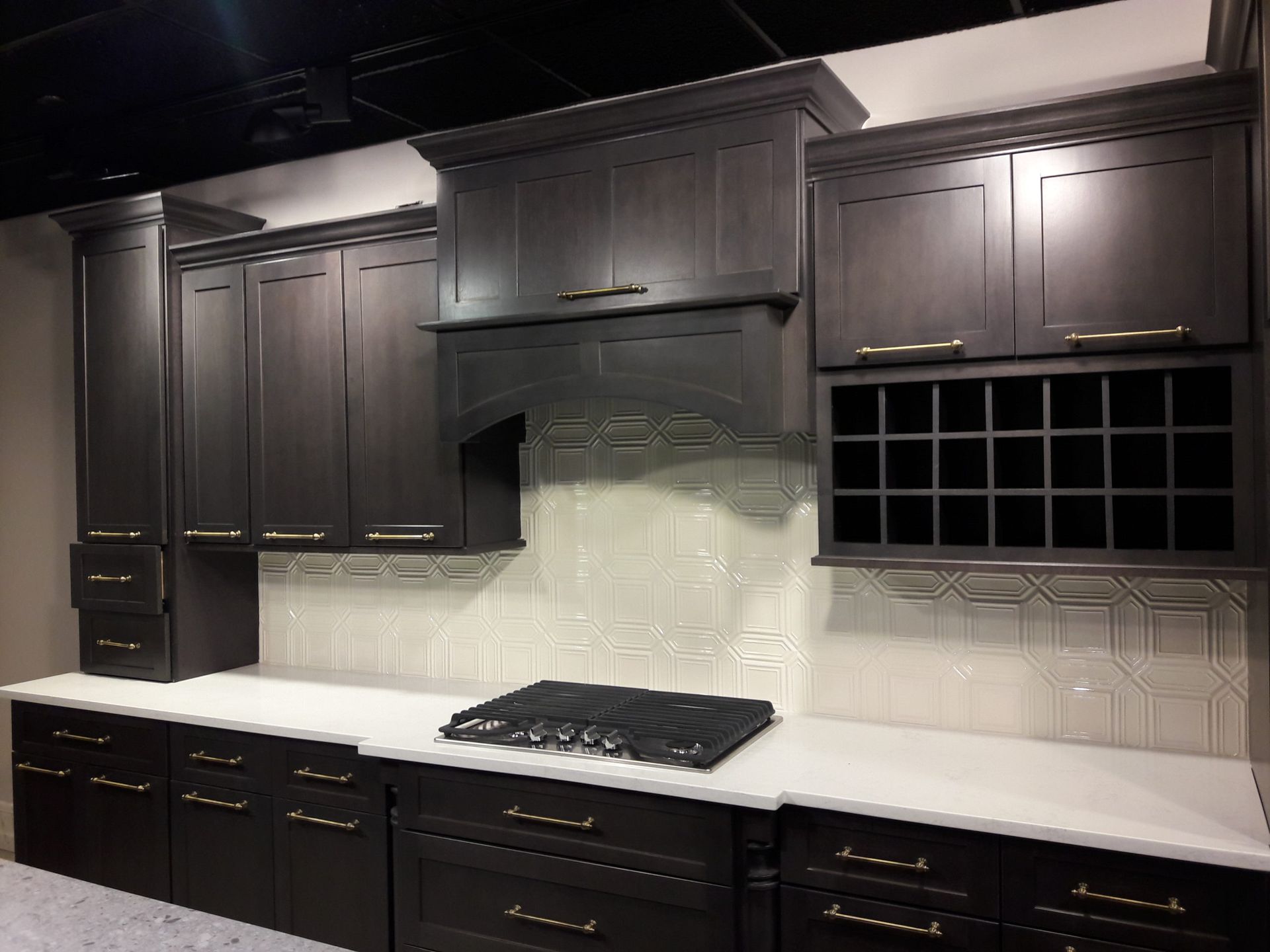 Dark gray kitchen cabinets with a white countertop and backsplash, a black stovetop, and wine rack.