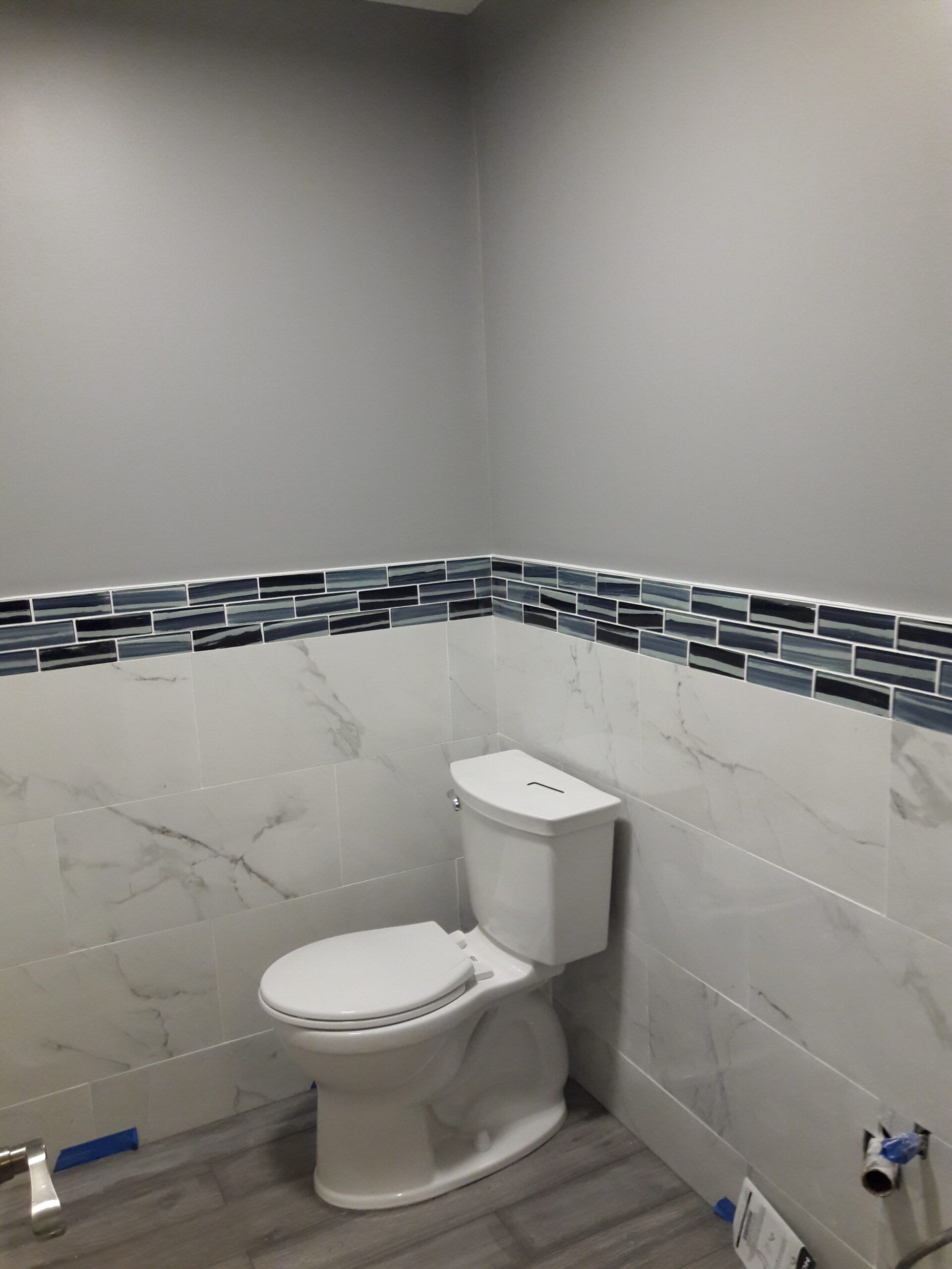 Bathroom with white toilet, white and grey marble tiles, and blue and black mosaic tile trim. Gray walls.