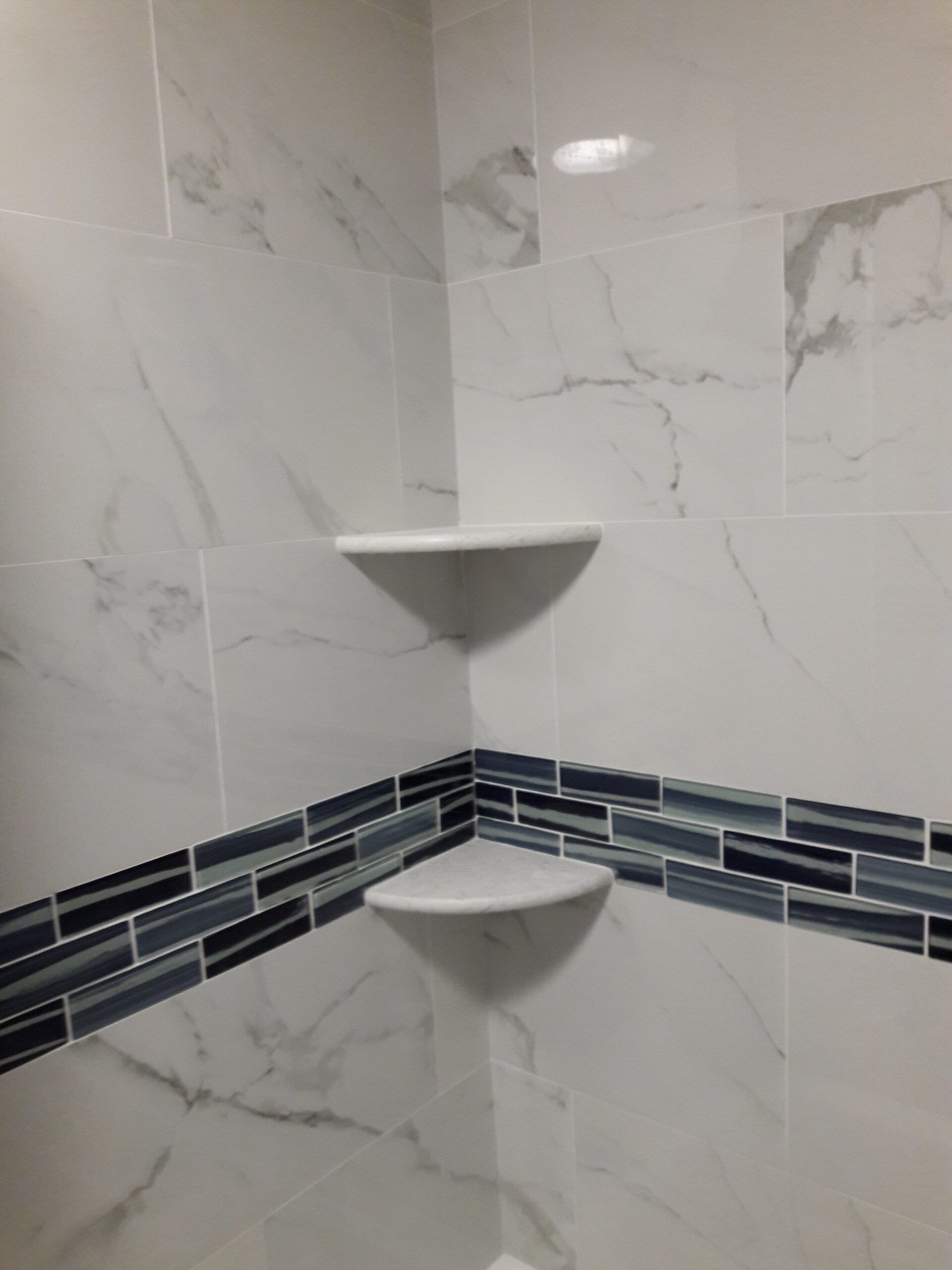 Corner shower with white marble-look tiles, two built-in corner shelves, and a blue mosaic tile accent.